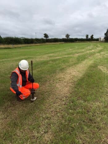 man wearing high visibility clothing and helmet kneeling down on grass with tool to investigate ground